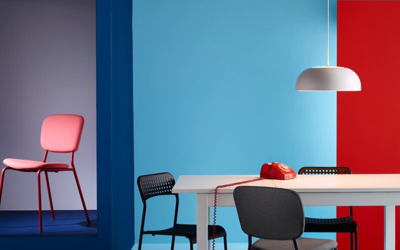 A modern room with a vibrant color scheme: a red chair in a blue alcove, a white table surrounded by black and gray chairs on the right, and a red retro phone on it. The room's stylish appeal is enhanced by custom-built wardrobes that seamlessly blend with the bold blue and red walls.